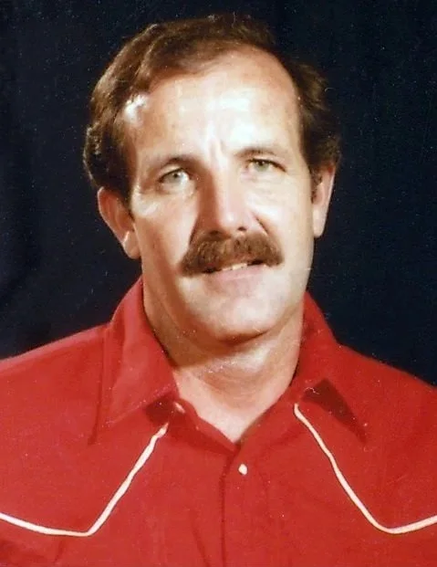 A man with a mustache and brown hair, wearing a red shirt with white piping on the shoulders, standing against a dark background.