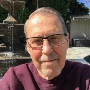 Close-up of a middle-aged man with glasses and a beard, smiling outdoors in front of a fountain.