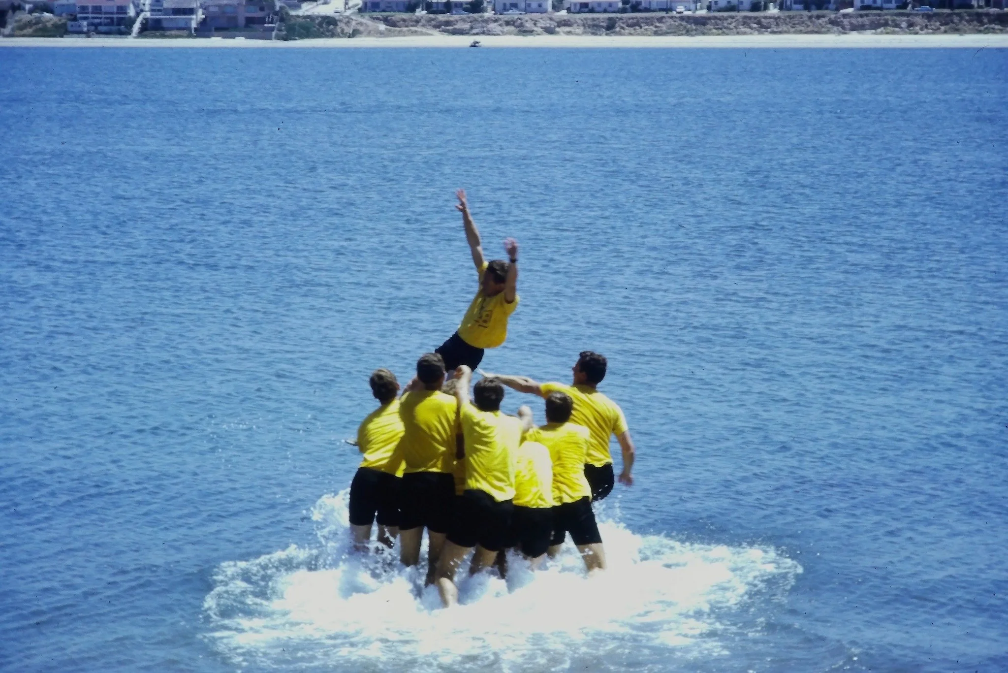 Group of people in yellow shirts engaging in a human pyramid or formation on a boat in a body of water, with one person standing on the shoulders of others in the middle.