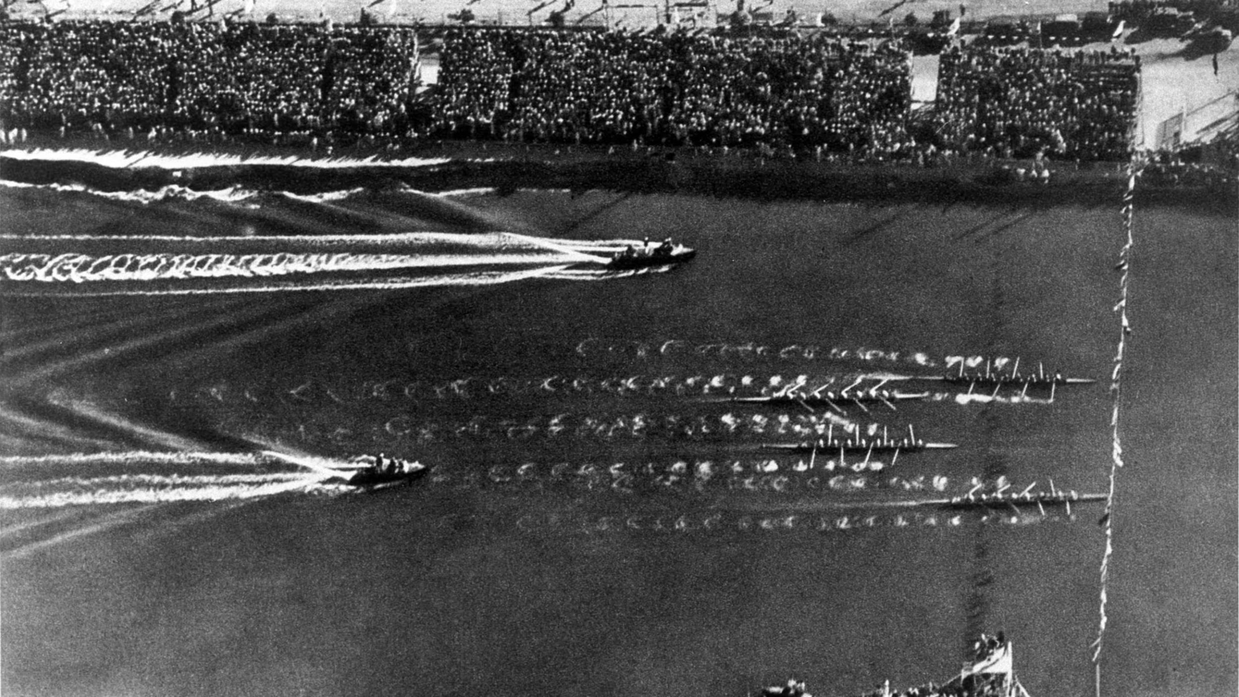 Historical black and white photograph of a military parade with boats or ships in the water, creating wakes as they move, and a large crowd of spectators behind a barrier along the shoreline.