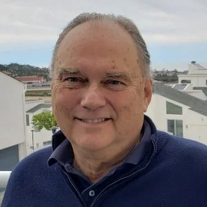 A smiling older man with gray hair, wearing a navy blue sweater and collared shirt, standing outdoors with buildings and trees in the background.