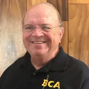 Smiling man wearing glasses and a black polo shirt with 'BCA' embroidered, standing in front of a wooden wall.