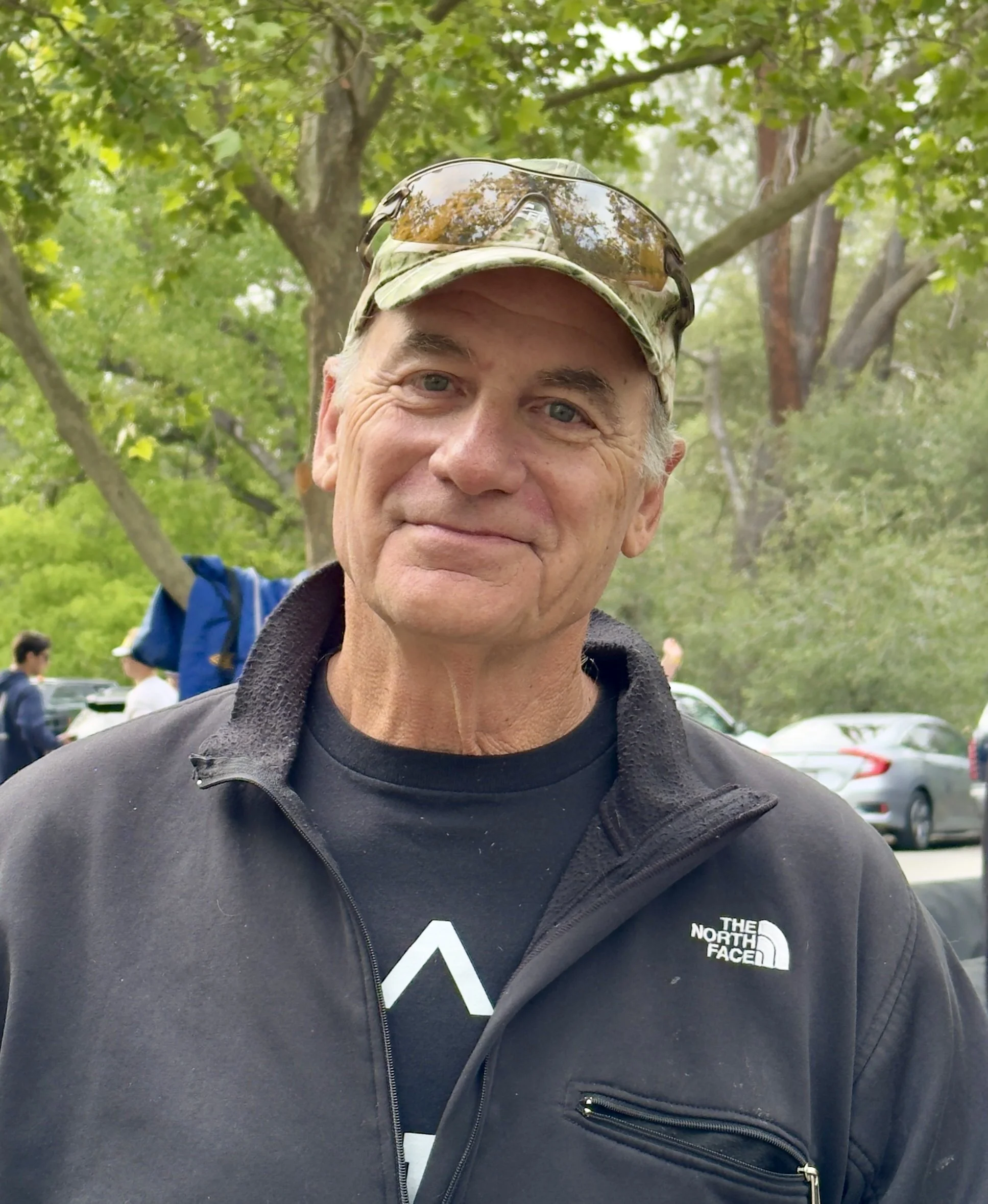 A man wearing a red and white cap with sunglasses on top, standing outdoors in a green, natural setting.