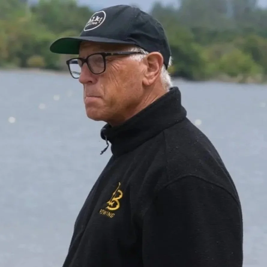 A man wearing glasses, a black cap, and a black jacket standing outdoors by a body of water with trees in the background.