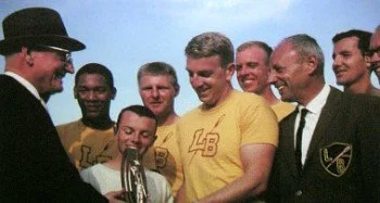 Group of men, including one in a yellow t-shirt with a racing logo, gathered outdoors, with one man wearing a black hat and sunglasses, smiling and looking at a small trophy.