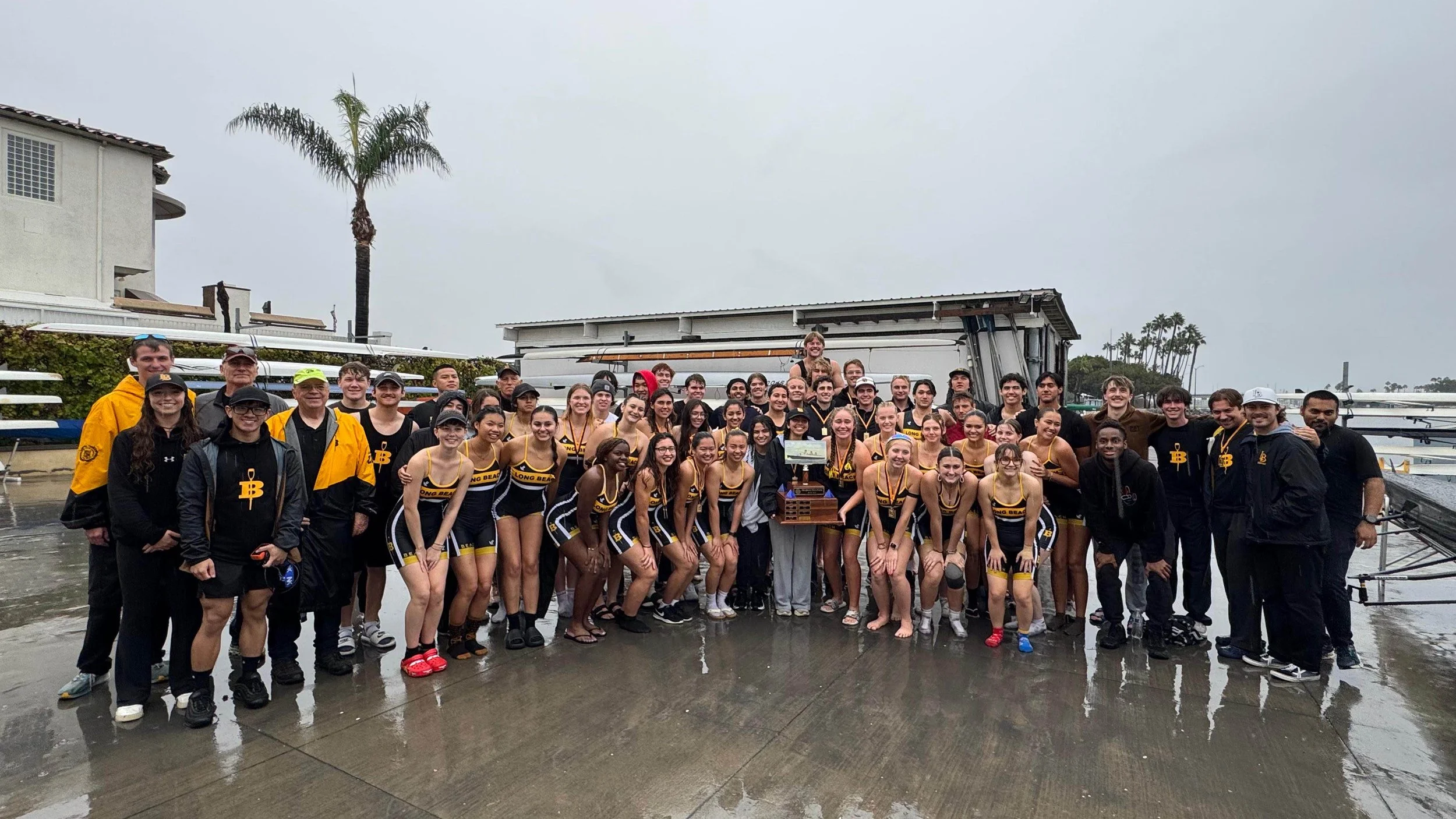 Group of young rowers and their coaches celebrating after a race, holding a trophy, on a rainy day at a dock with rowing shells in the background.