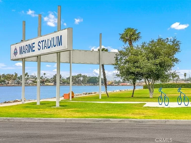 Large sign reading 'Marine Stadium' near water with trees, grass, and two bicycles parked on the right side under a partial blue sky.
