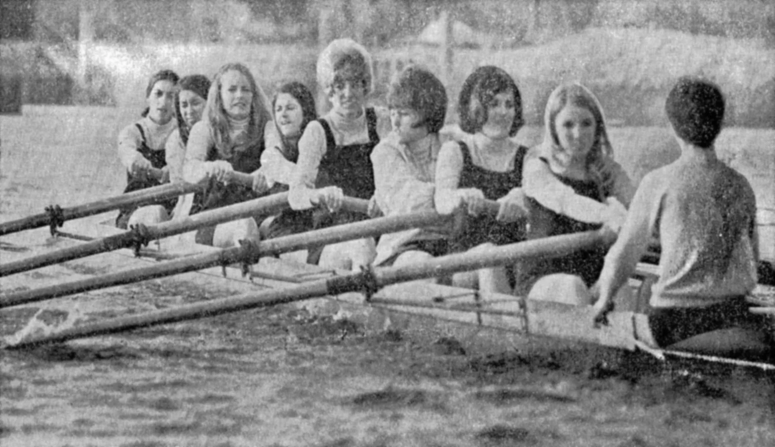 A vintage black and white photo of seven young girls and one boy sitting on a rowboat tied to a dock or shore, with a grassy field in the background.