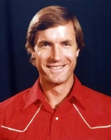 A smiling man with brown hair wearing a red western-style shirt.