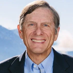 A middle-aged man smiling outdoors with mountains and blue sky in the background.