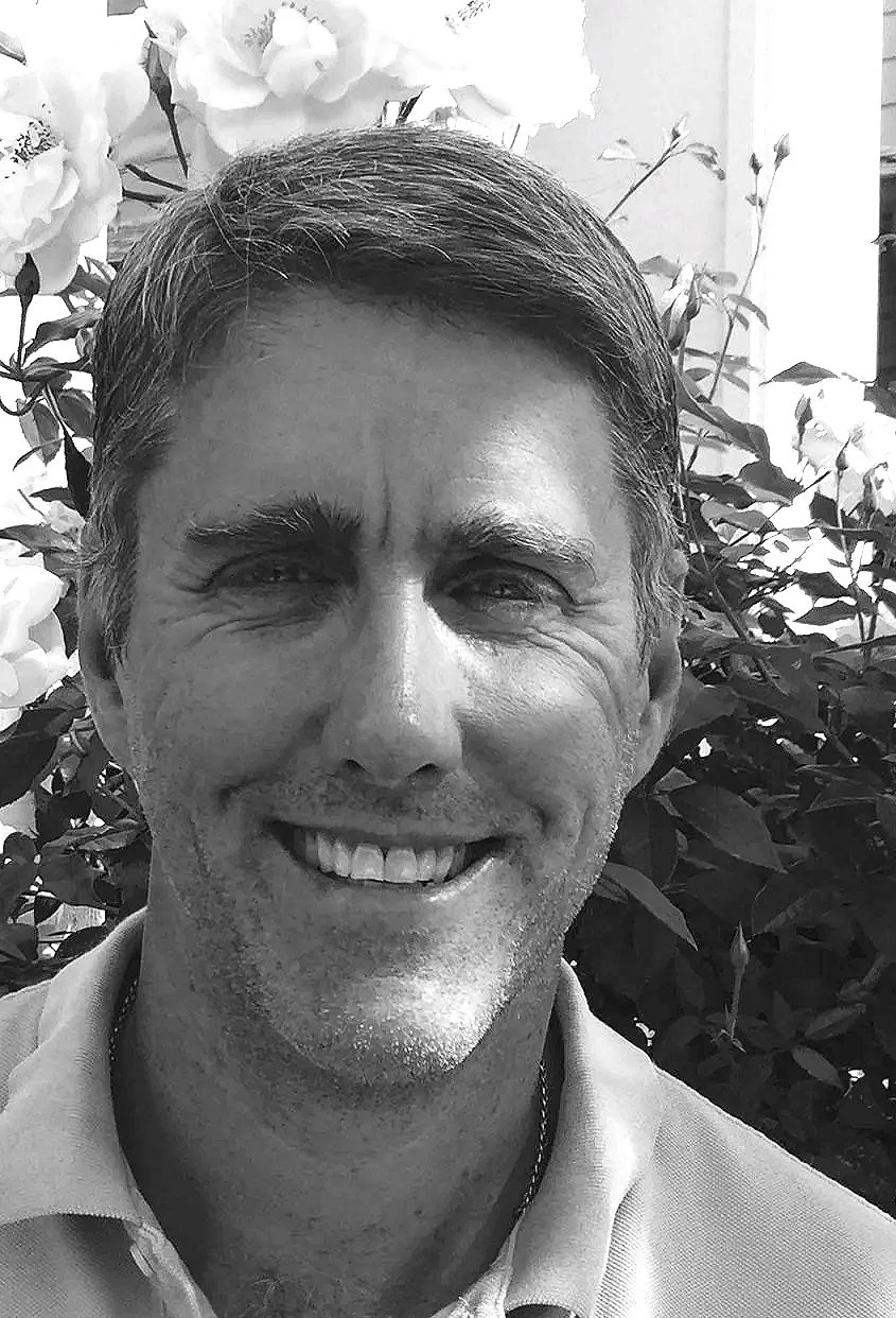 A smiling man with short hair, standing in front of blooming flowers. The photo is in black and white.