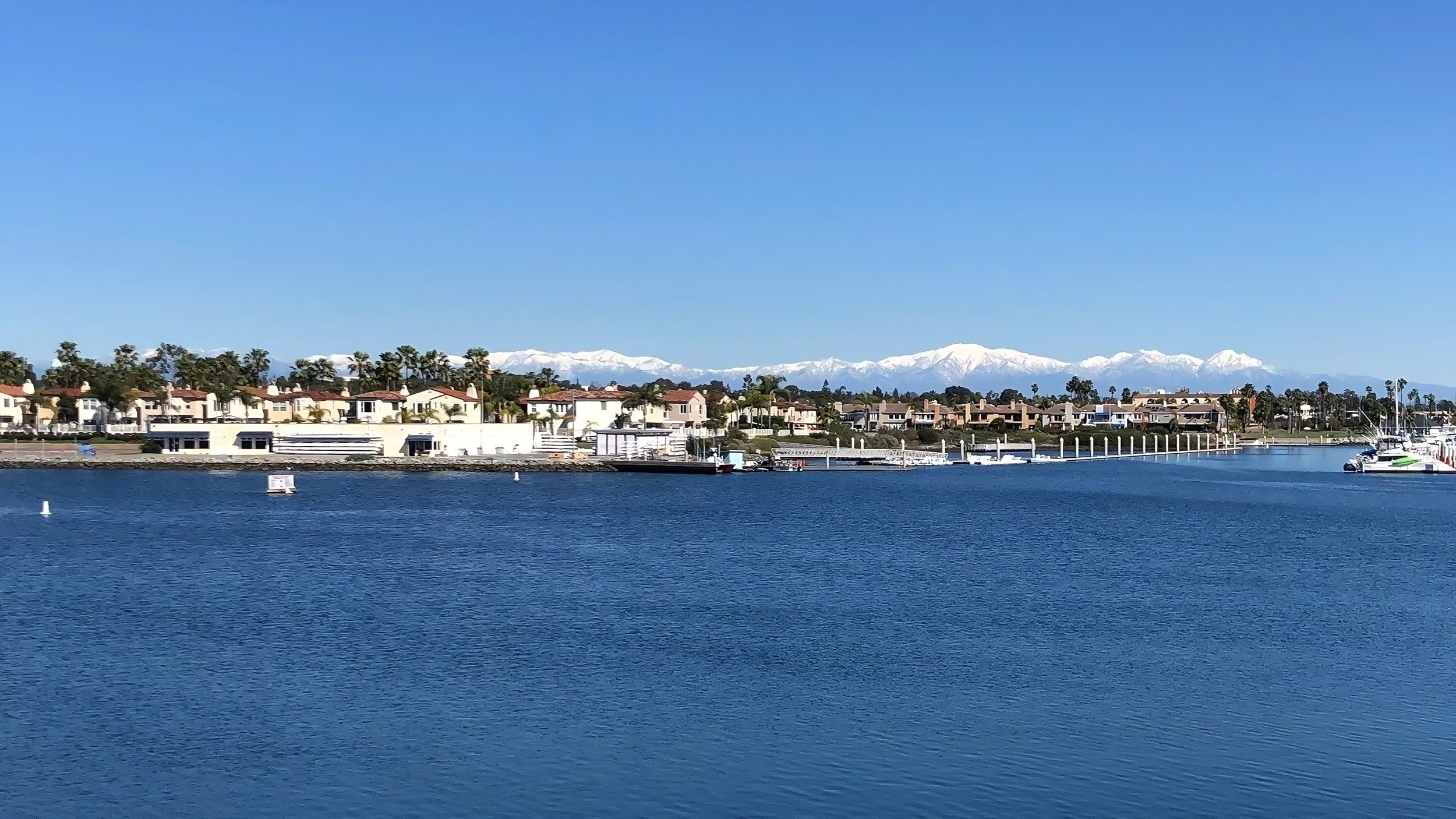 View of a calm harbor with boats docked, residential houses along the shore, palm trees, and snow-capped mountains in the background under a clear blue sky.