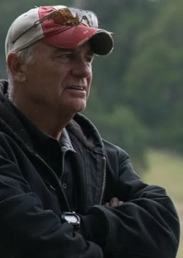 A man wearing a baseball cap and jacket, standing outdoors with crossed arms and a serious expression.