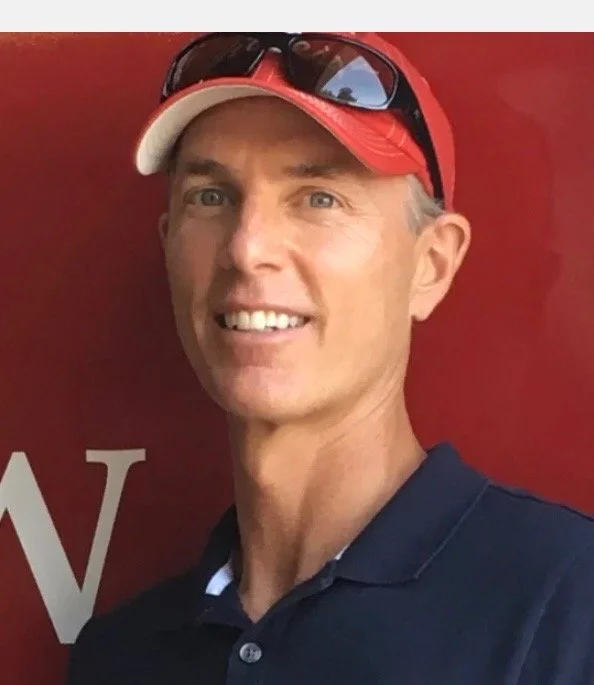 A smiling man wearing a red baseball cap, sunglasses on top, and a navy blue polo shirt, standing in front of a red background.