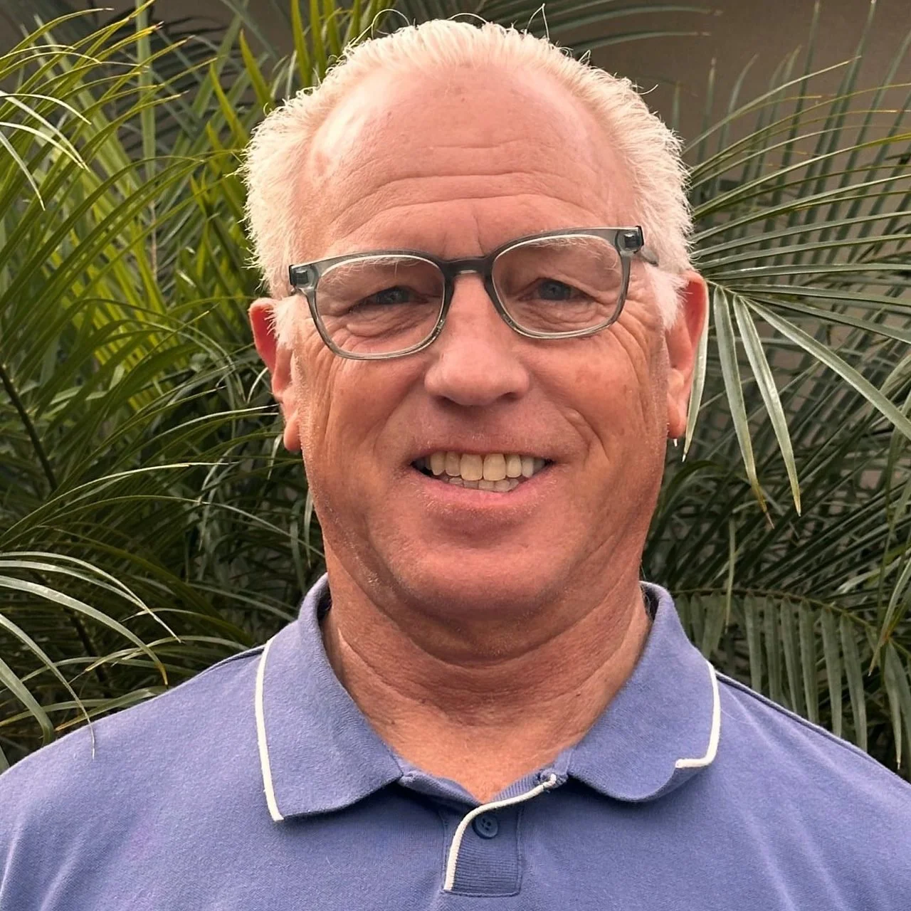 A smiling man with glasses and white hair standing in front of green plants, wearing a blue polo shirt.