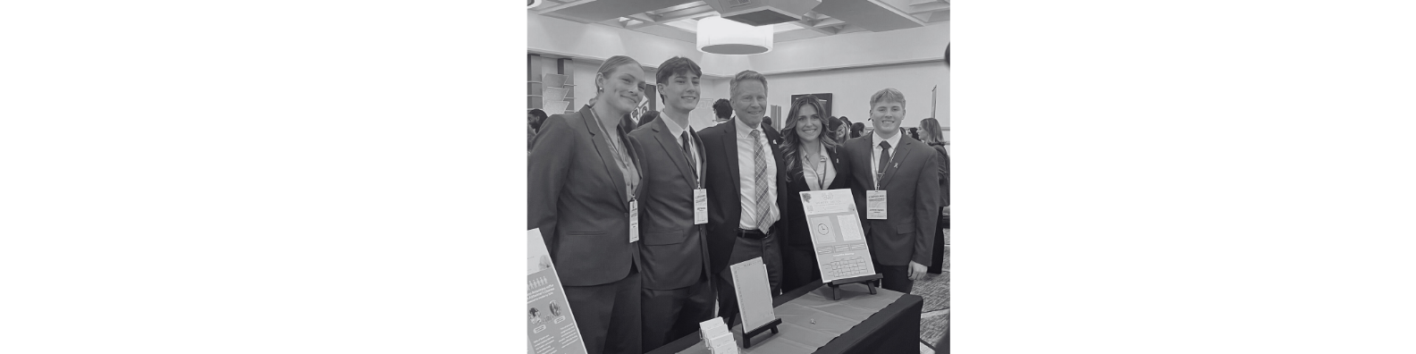 Group of five people in business attire standing at a conference booth with informational displays and promotional material on the table.