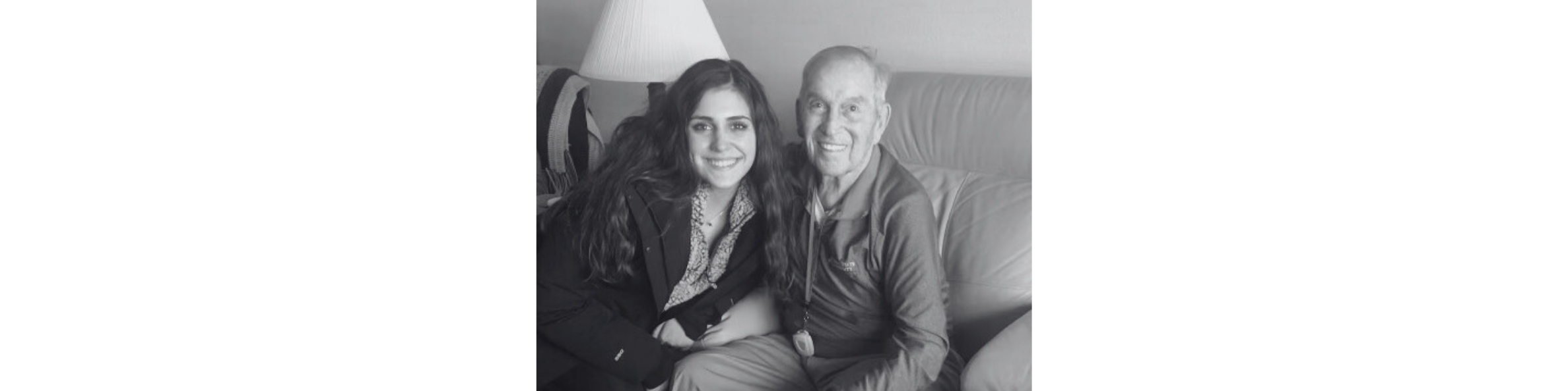 A young woman and an elderly man sit together on a couch, smiling at the camera. The woman has long, dark hair and is wearing a dark jacket, while the man has short, light hair and is wearing a long-sleeve shirt. There is a lamp in the background.