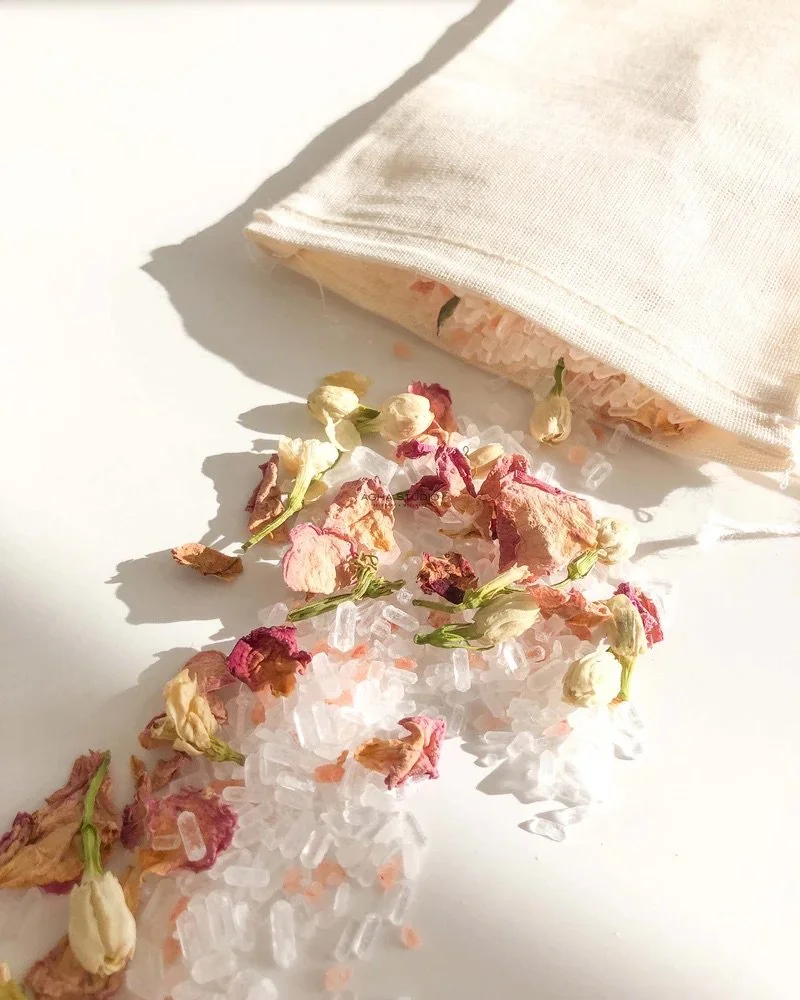 Dried pink and white flowers alongside pink Himalayan salt crystals spilling from a beige fabric bag onto a white surface.