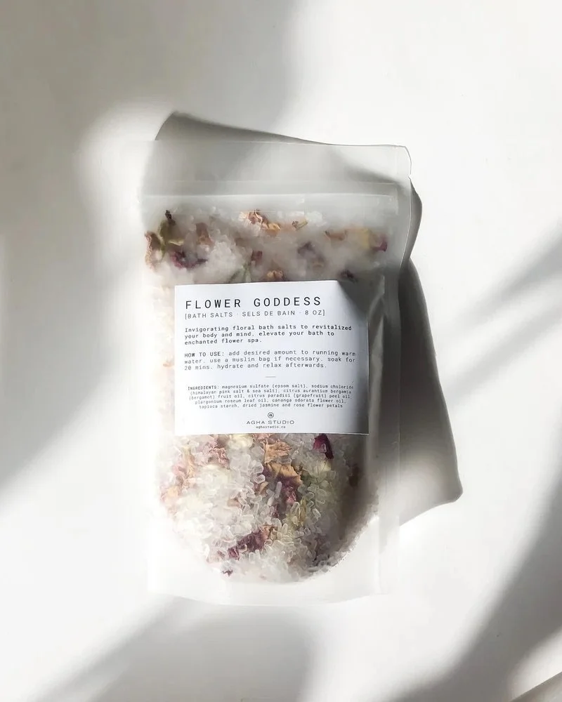 A clear plastic bag of bath salts called 'Flower Goddess' with dried flower petals inside, lying on a white surface.