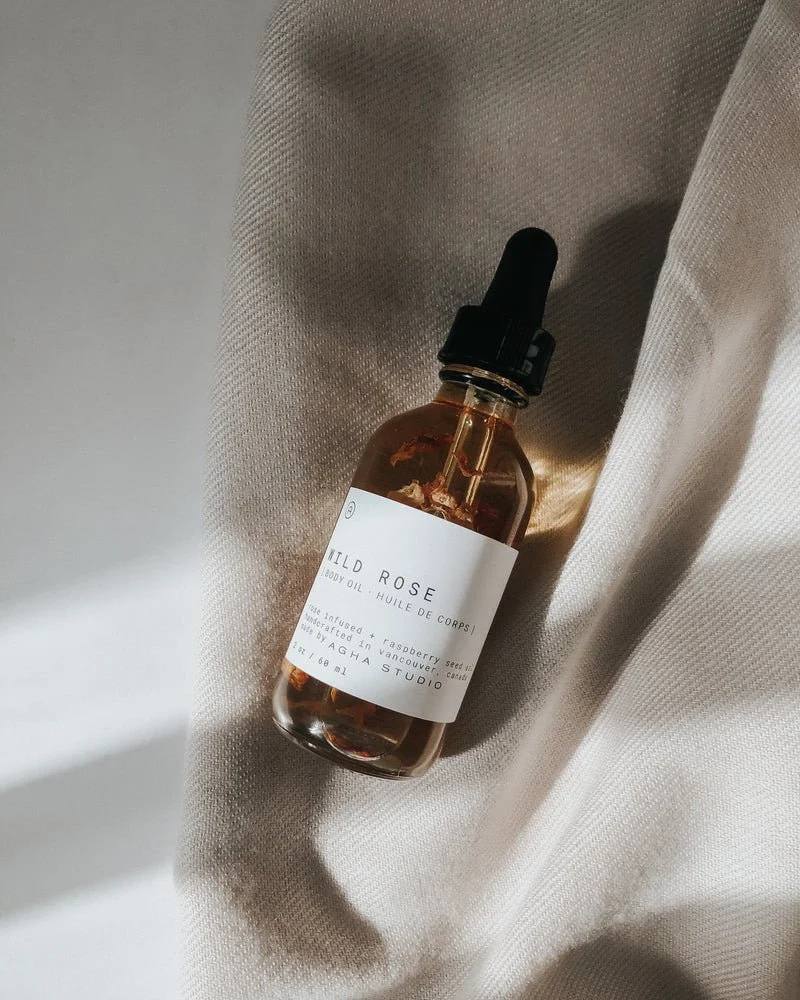 A small amber glass bottle of Wild Rose body oil with a black dropper cap resting on cream-colored fabric.