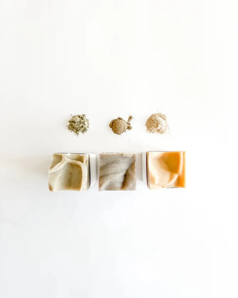 Three bars of soap with different colors and three piles of powder or clay above them on a white background