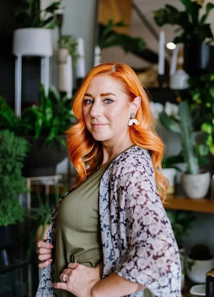 A woman with long red hair styled in loose waves, wearing a green top, a patterned kimono, and white earrings, standing in a room with various potted plants and modern decor.