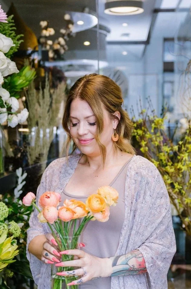 A woman with tattoos on her arm holding a bouquet of pink and orange flowers in a floral shop.