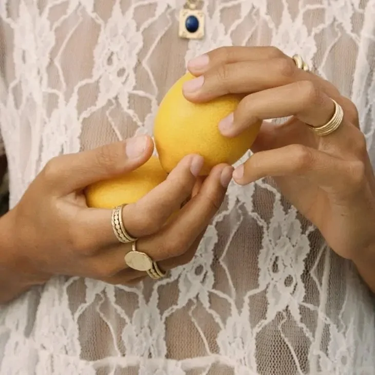Hands with rings holding a yellow lemon, with a lace top in the background.
