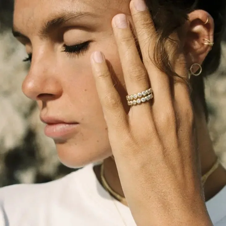 A woman touching her face with her left hand, showing a pearl ring on her ring finger and a gold hoop earring, with her eyes closed and wearing minimal makeup.