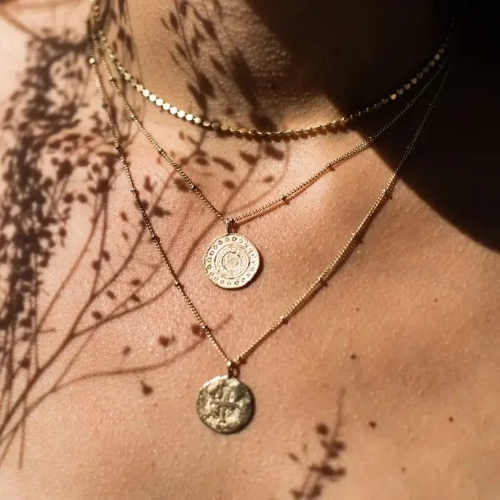 A close-up of a person's neck and shoulder showing layered gold necklaces with coin pendants, casting shadows on the skin.
