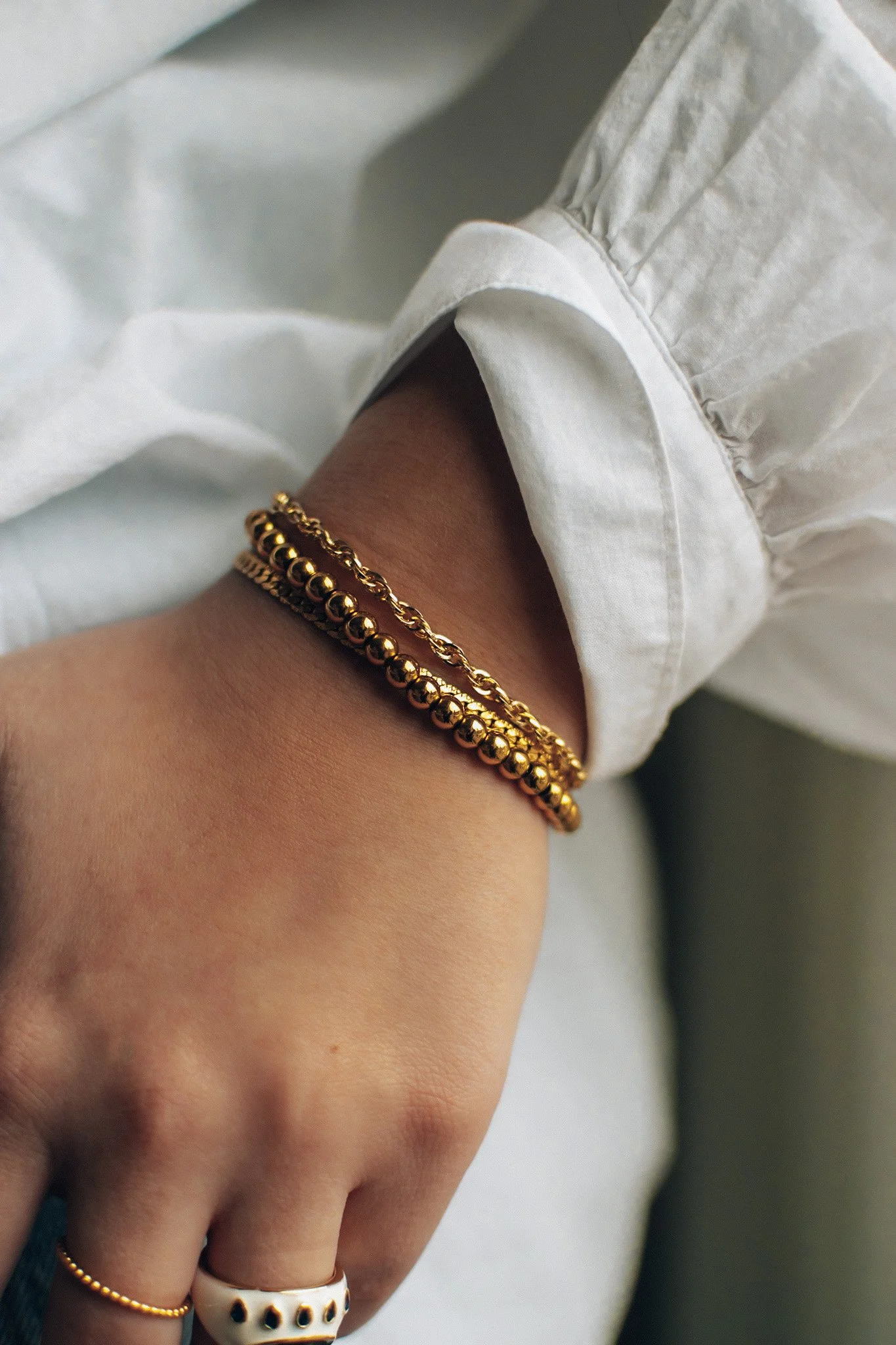 Close-up of a hand with a white shirt sleeve pushed up, wearing gold jewelry including bracelets and rings.