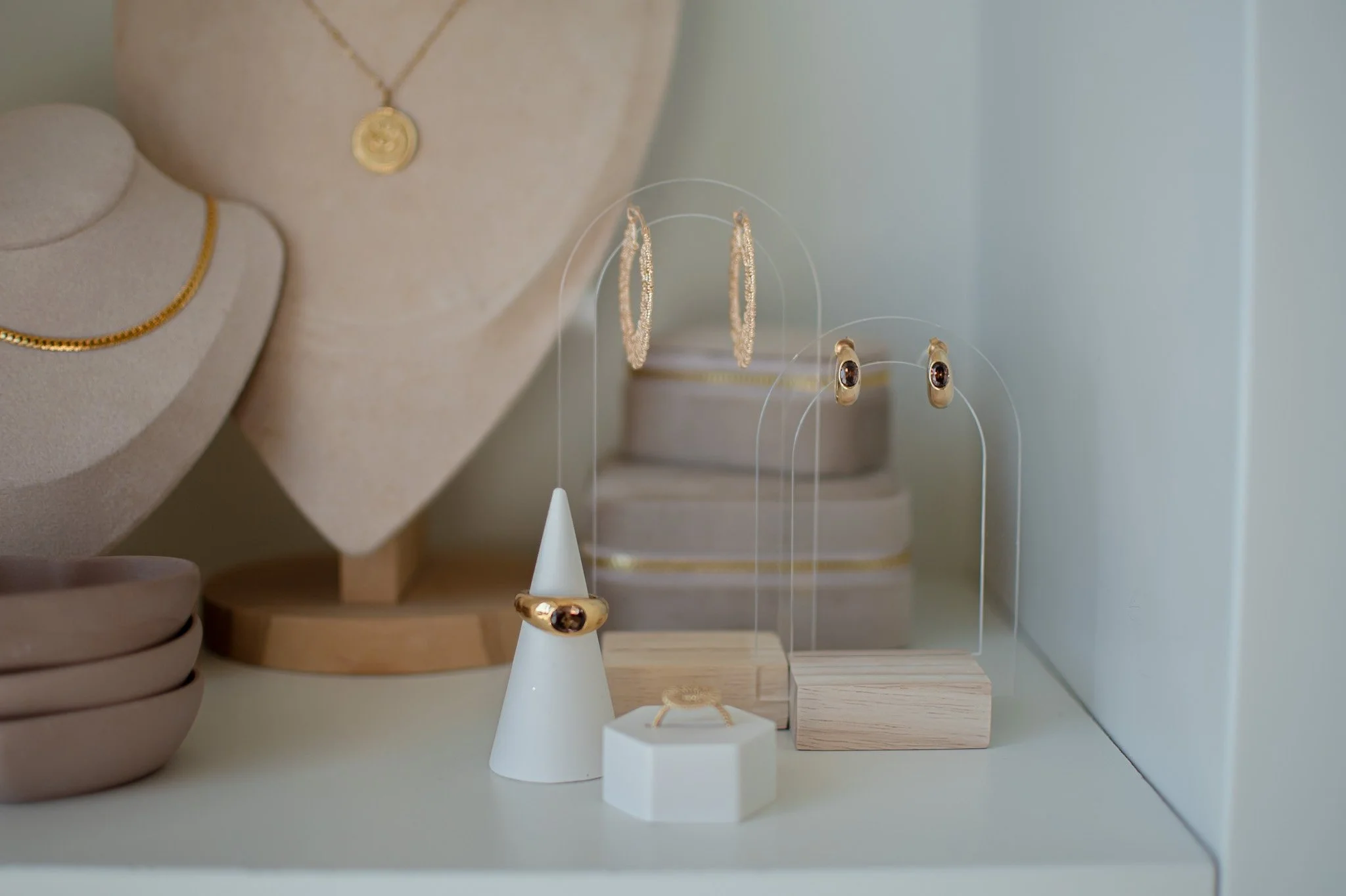 Display of gold jewelry including rings, hoop earrings, and a necklace on white stands and a beige ring holder, arranged on a wooden shelf.