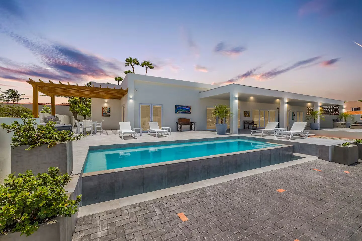 Modern backyard with a swimming pool, white lounge chairs, and a white house with outdoor seating and potted plants at sunset.