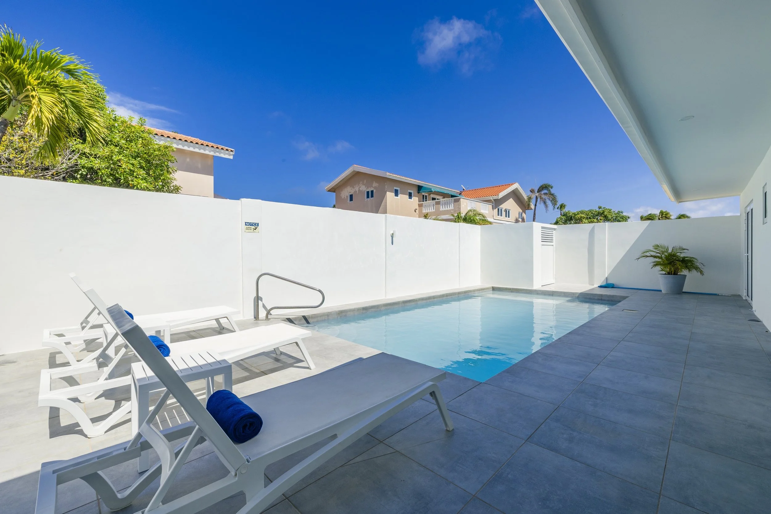 View of a private backyard swimming pool with white lounge chairs, blue towels, potted plants, a white wall, and neighboring houses under a blue sky.