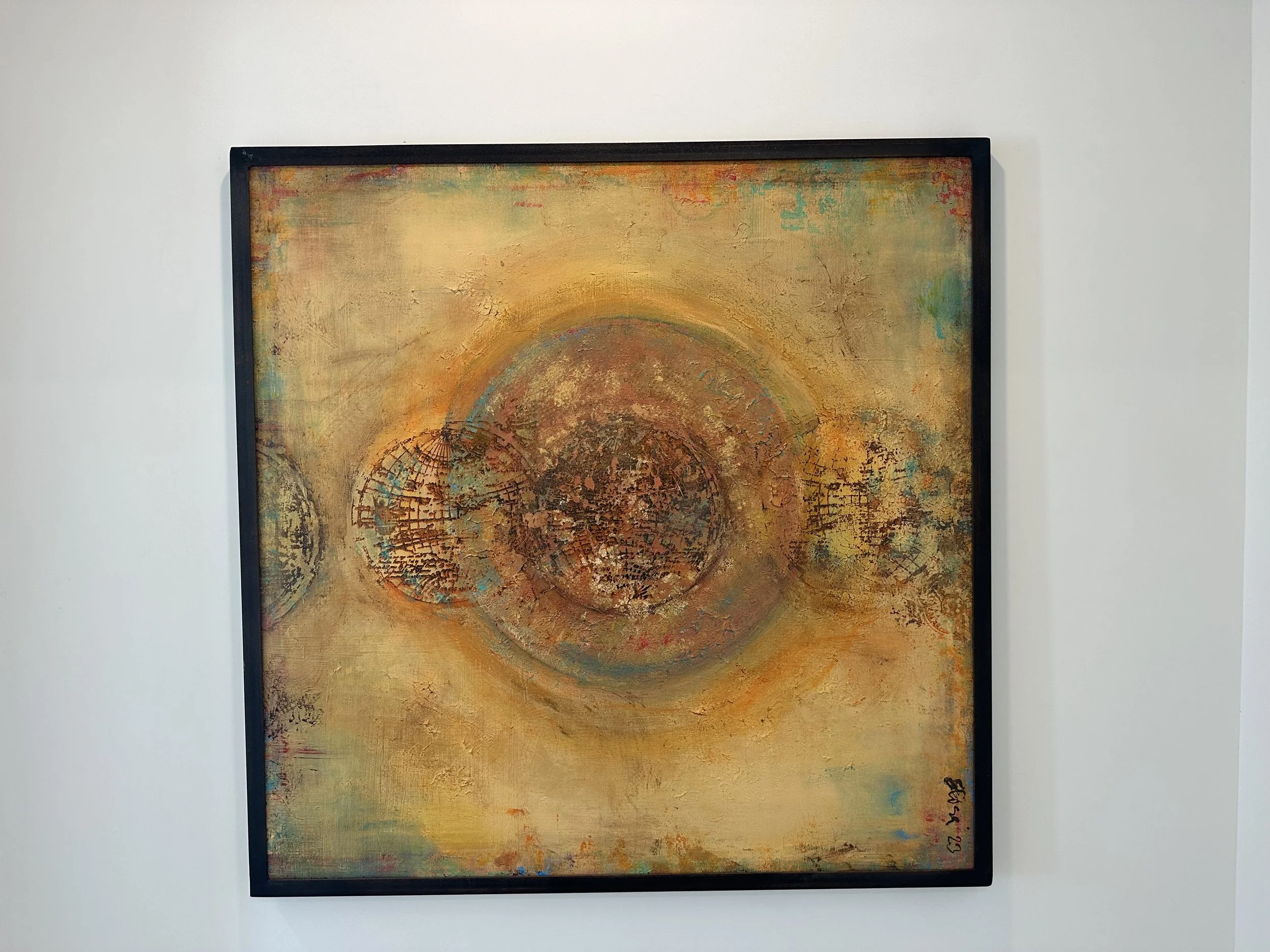 Abstract painting with orange, yellow, brown, and blue hues, featuring overlapping circular shapes with textured patterns, framed in black on a white wall.
