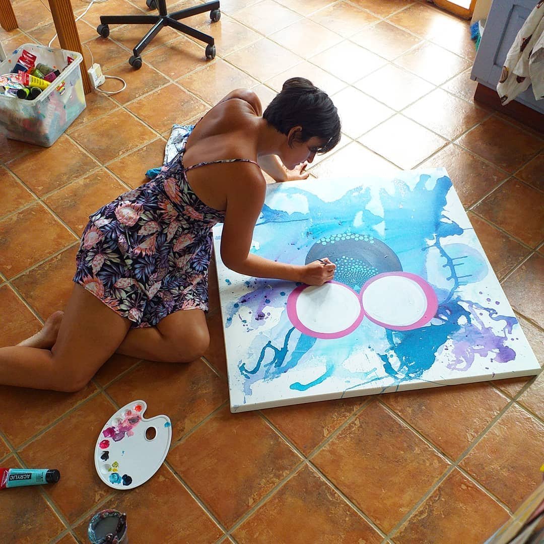 A woman in a floral dress is kneeling on a tiled floor, painting on a large canvas with abstract blue and purple splashes and two large circular shapes outlined in pink. Art supplies are nearby, including a palette with paint and a tube of paint.