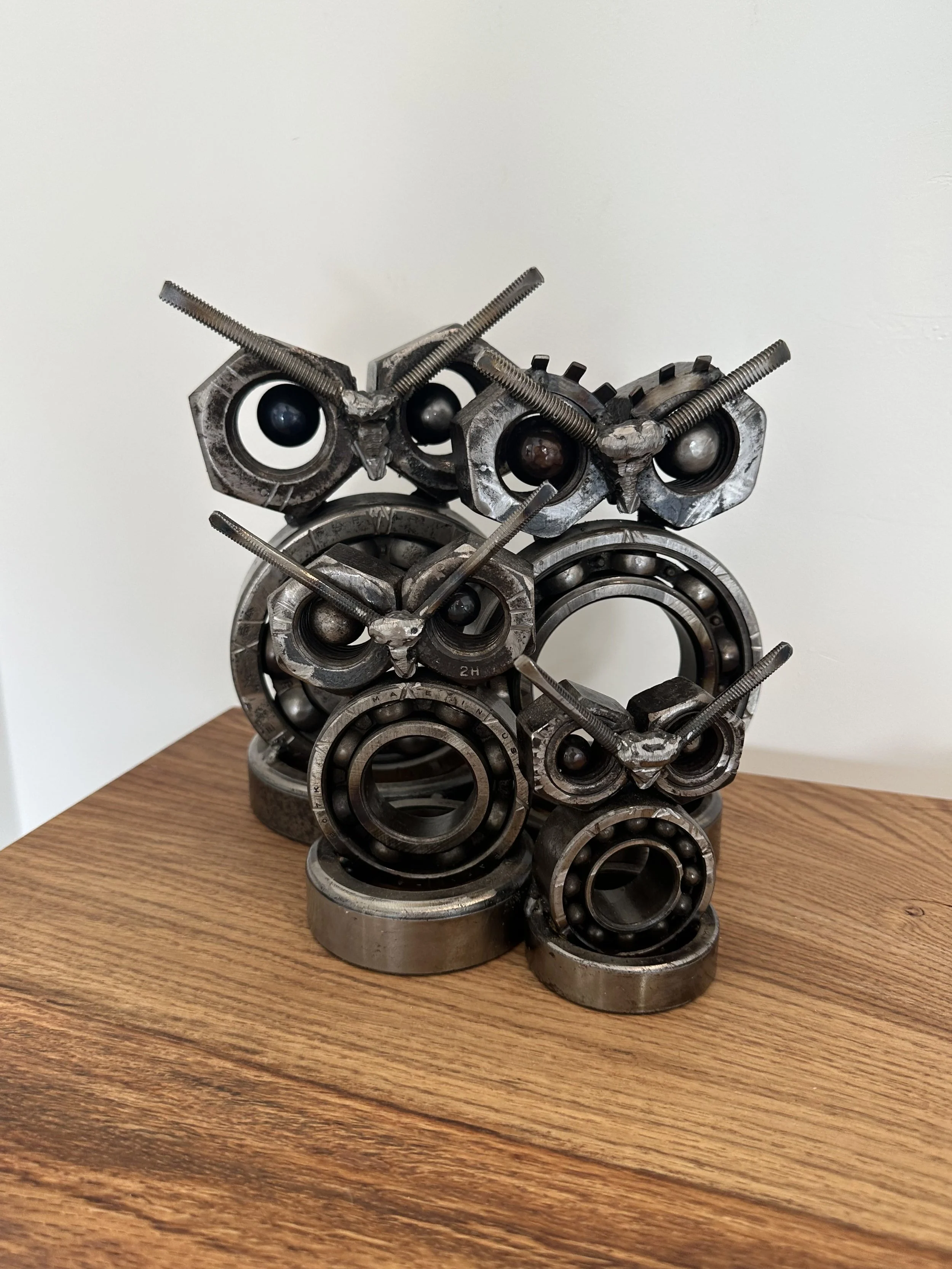 An owl sculpture made of various metal parts including ball bearings, bolts, and bicycle gears, arranged to resemble an owl on a wooden surface.