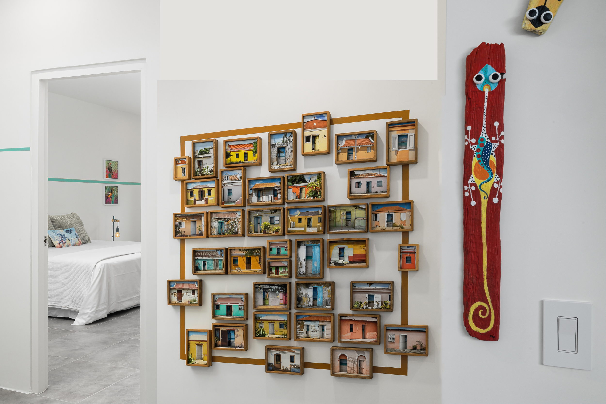 Wall-mounted photo display featuring numerous small framed images of colorful houses, adjacent to a painted wooden giraffe with googly eyes on the wall, and a glimpse of a decorated bedroom with pillows and artwork in the background.