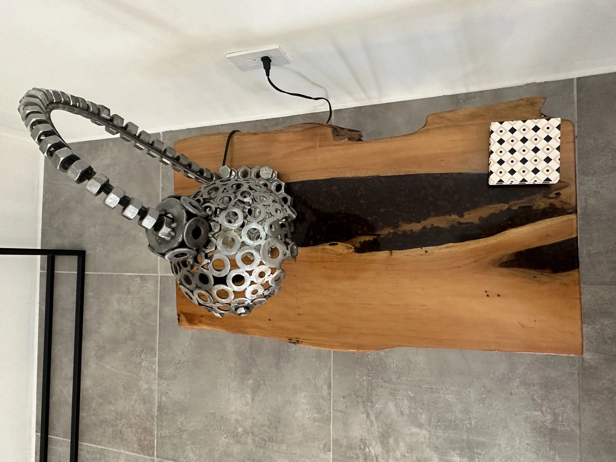 A decorative lamp made from metal washers and bolts, placed on a wooden slab with a small patterned tile nearby.