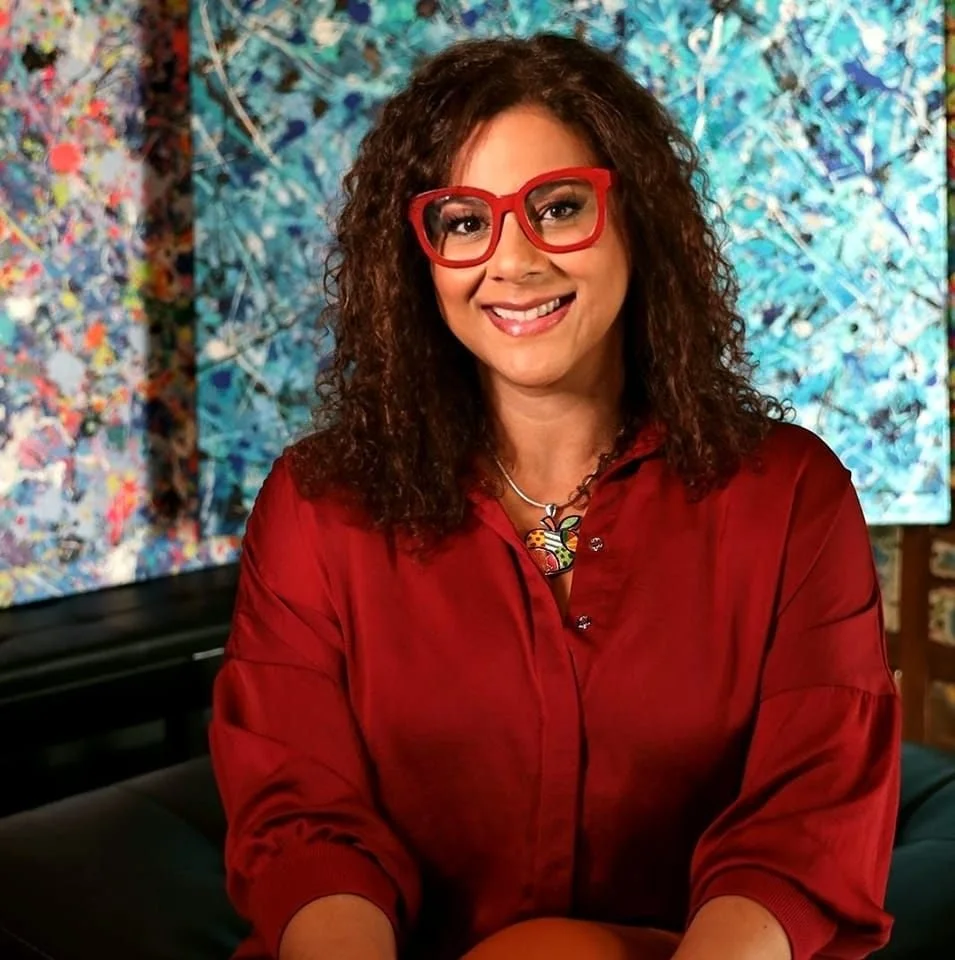 A woman with curly brown hair and red glasses smiling at the camera, wearing a red blouse, with colorful abstract paintings in the background.