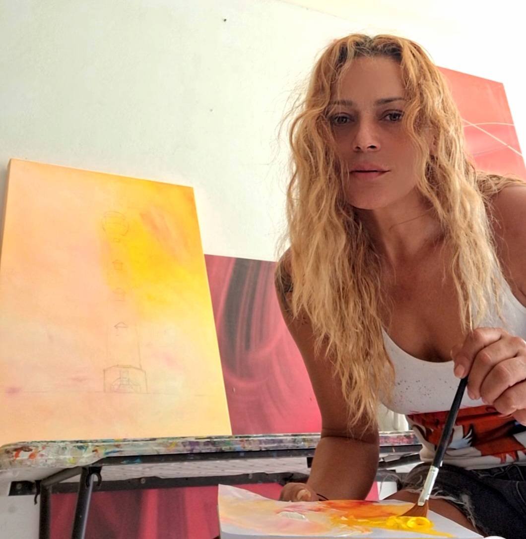 A woman with curly blonde hair painting on a canvas with yellow and orange colors in an art studio.