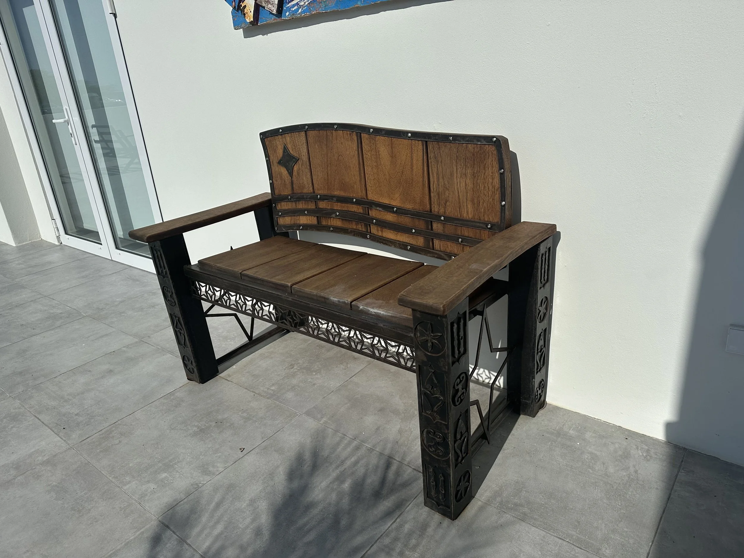 A wooden and metal garden bench with decorative details, placed on a tiled patio outside a building with a white wall and sliding glass door.