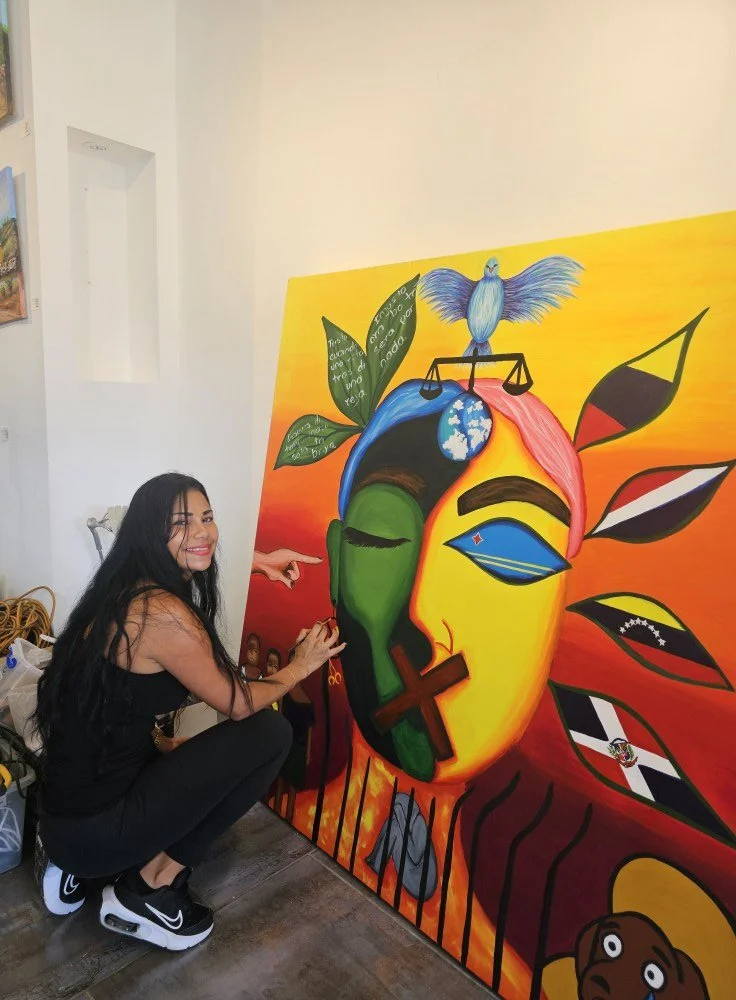Woman with long black hair squatting and smiling next to a colorful, abstract mural of a face with various symbols and flags on it, set indoors.