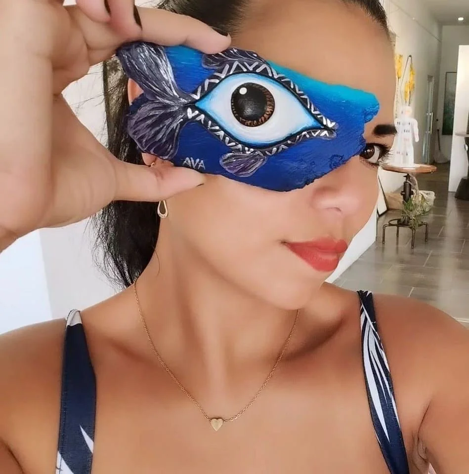 A woman with red lipstick holding a blue eye-shaped object with a fish design over her right eye, inside a room with modern decor.