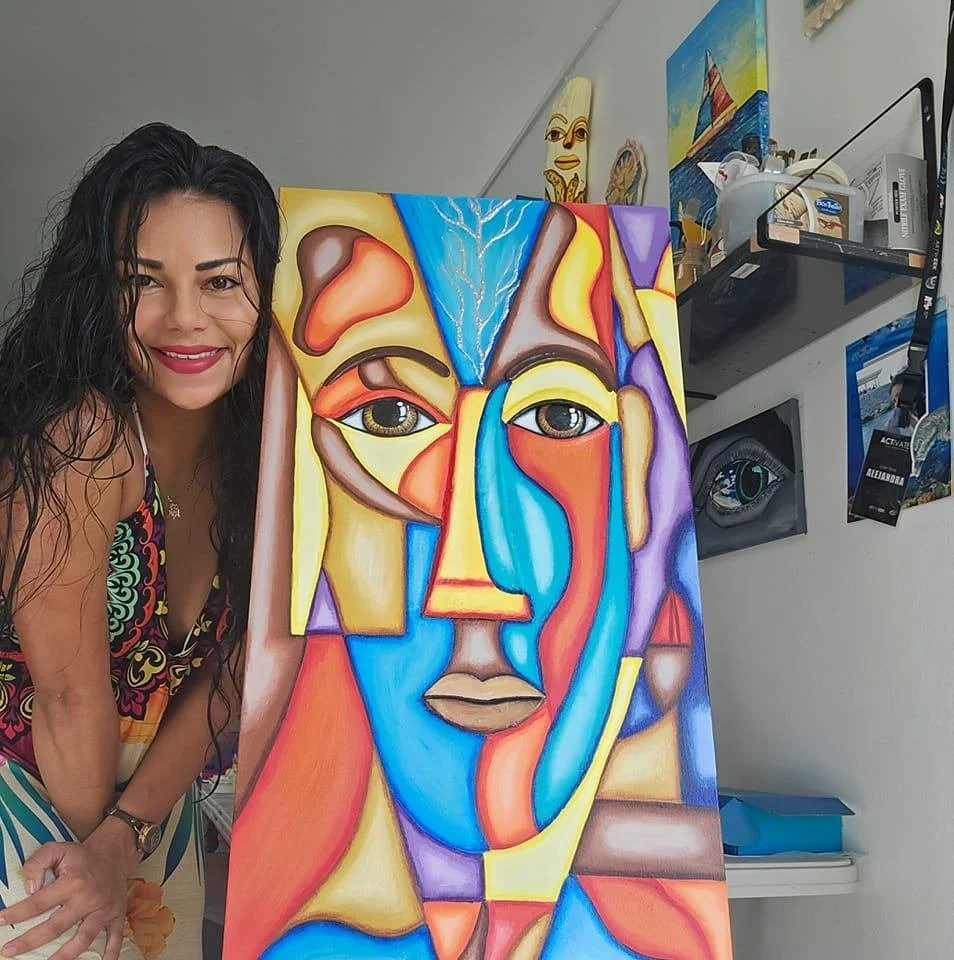 A woman with dark curly hair smiling next to a colorful abstract painting of a face.