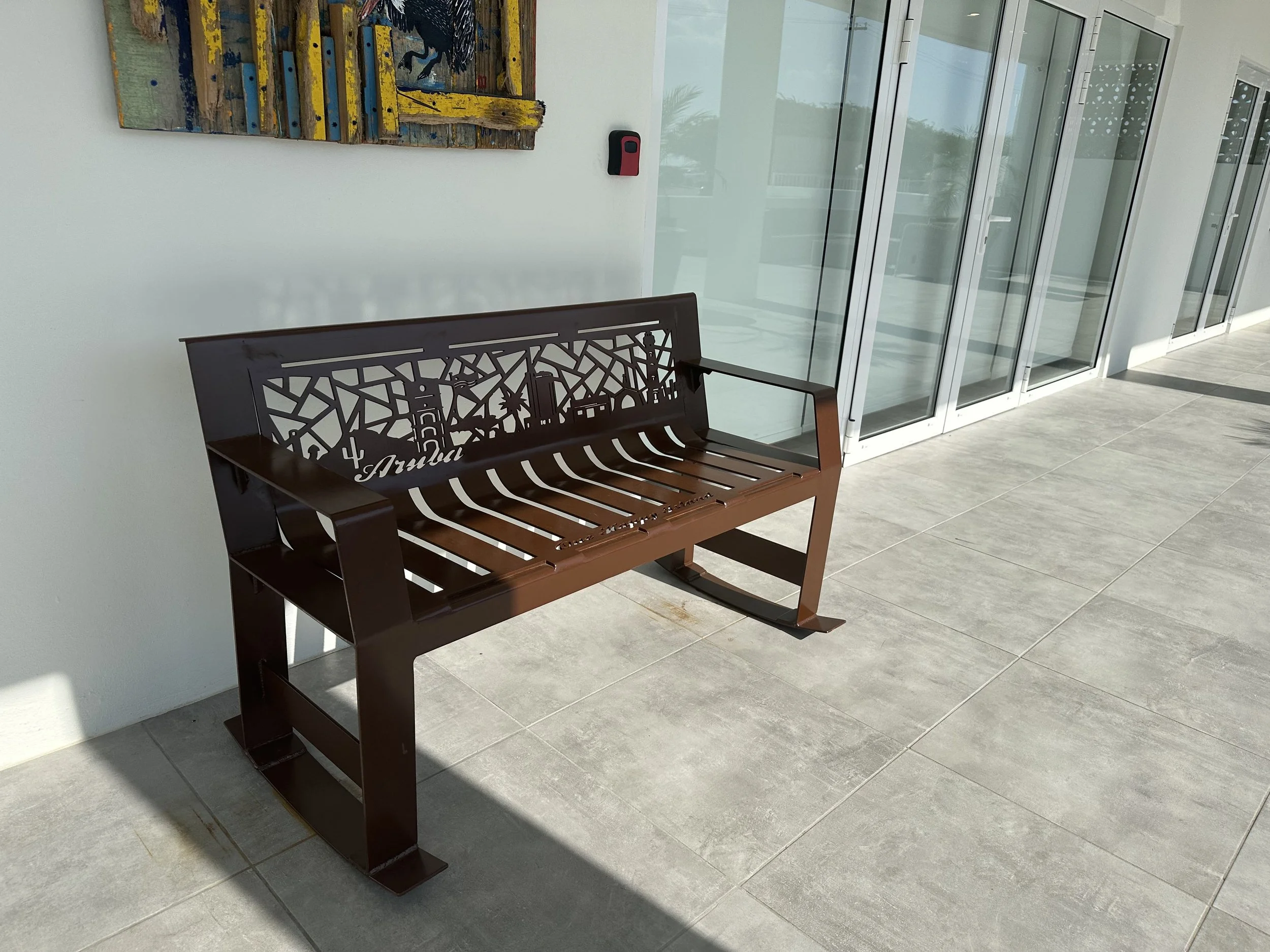 Brown metal bench with cutout cityscape and lighthouse design, positioned on a gray tiled outdoor patio near glass sliding doors, with colorful painting on the wall behind.