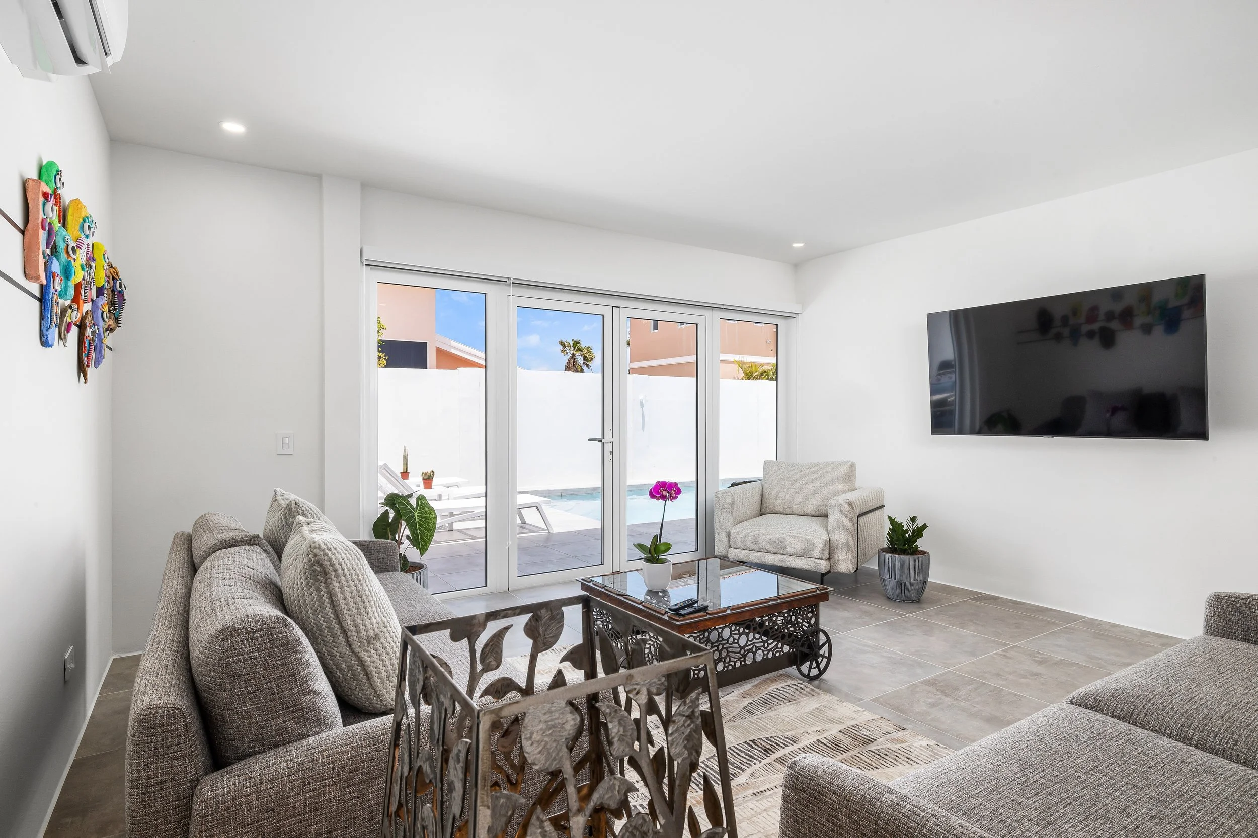 Modern living room with beige sofas, a white armchair, a glass coffee table with a potted orchid, plants, a wall-mounted TV, and large sliding glass doors leading to an outdoor patio with pool.