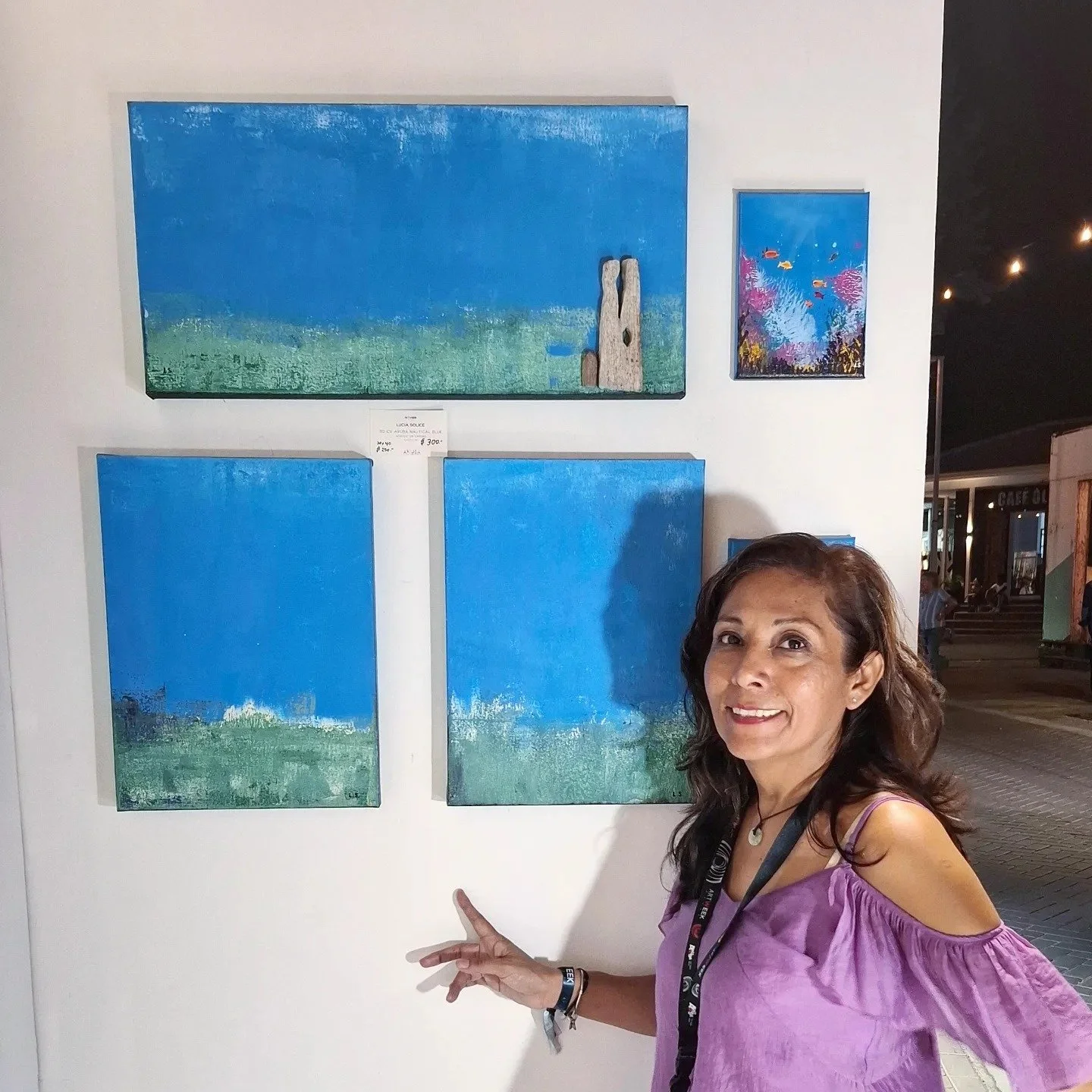 A woman wearing a pink blouse and a lanyard points at a collection of four paintings on a white wall, featuring abstract blue and green landscapes, with a nighttime outdoor scene in the background.