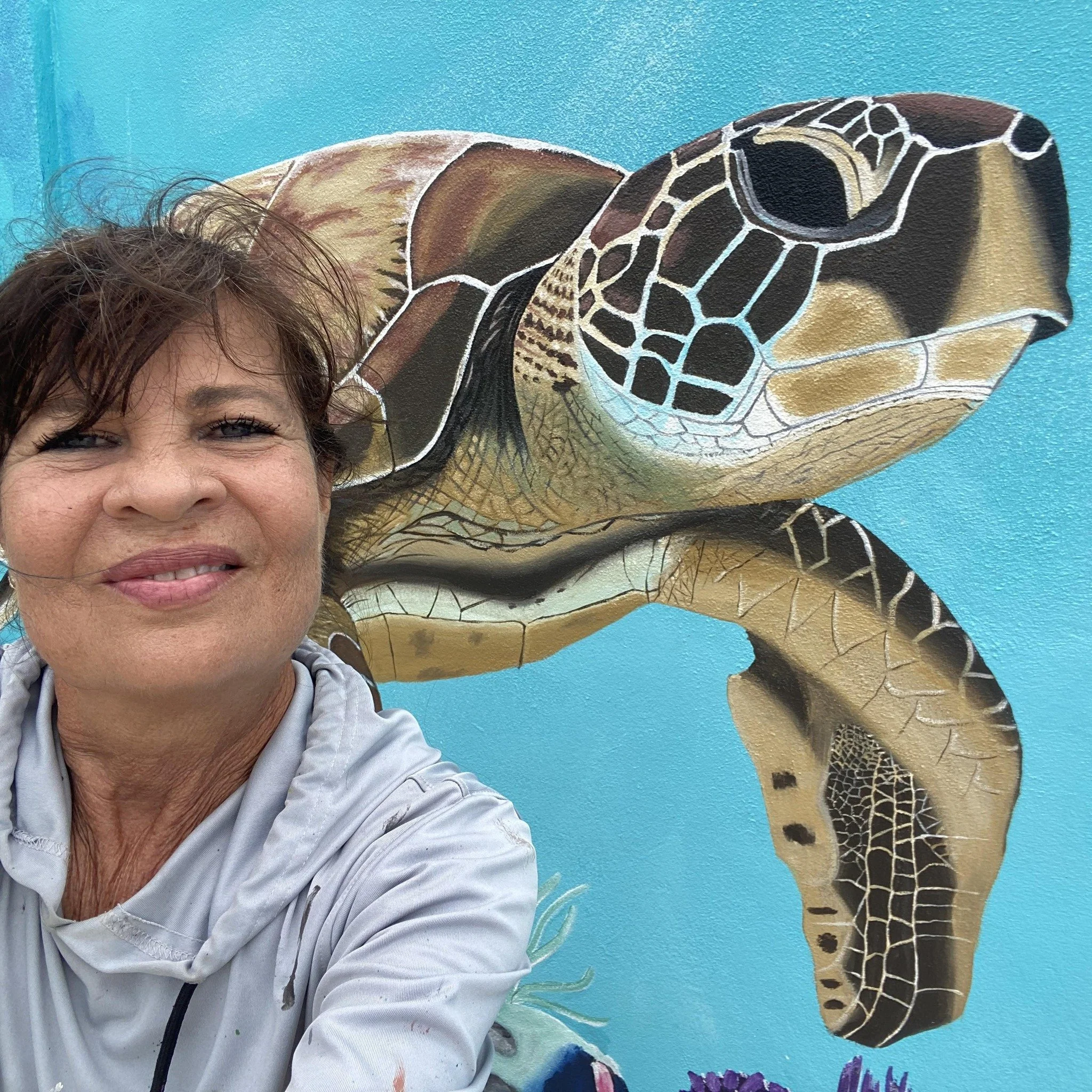 A woman taking a selfie in front of a mural of a sea turtle on a bright blue wall.