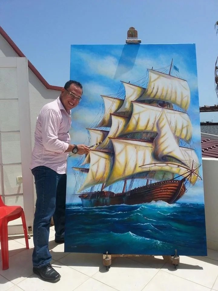 A man standing next to a large, colorful painting of a sailing ship on the water, pointing at it with a smile.